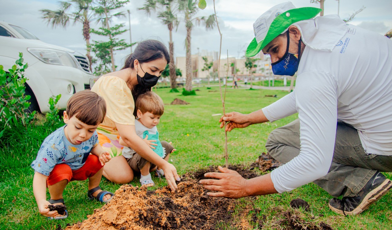 uma mulher e duas crianças plantam uma muda, auxiliados por técnico da prefeitura